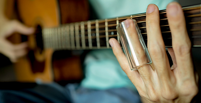 Close up of guitar player playing acoustic guitar with a glass guitar slide on his ring finger.